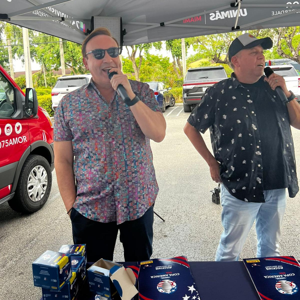 Two men speaking into microphones under a tent, with promotional materials on a table.