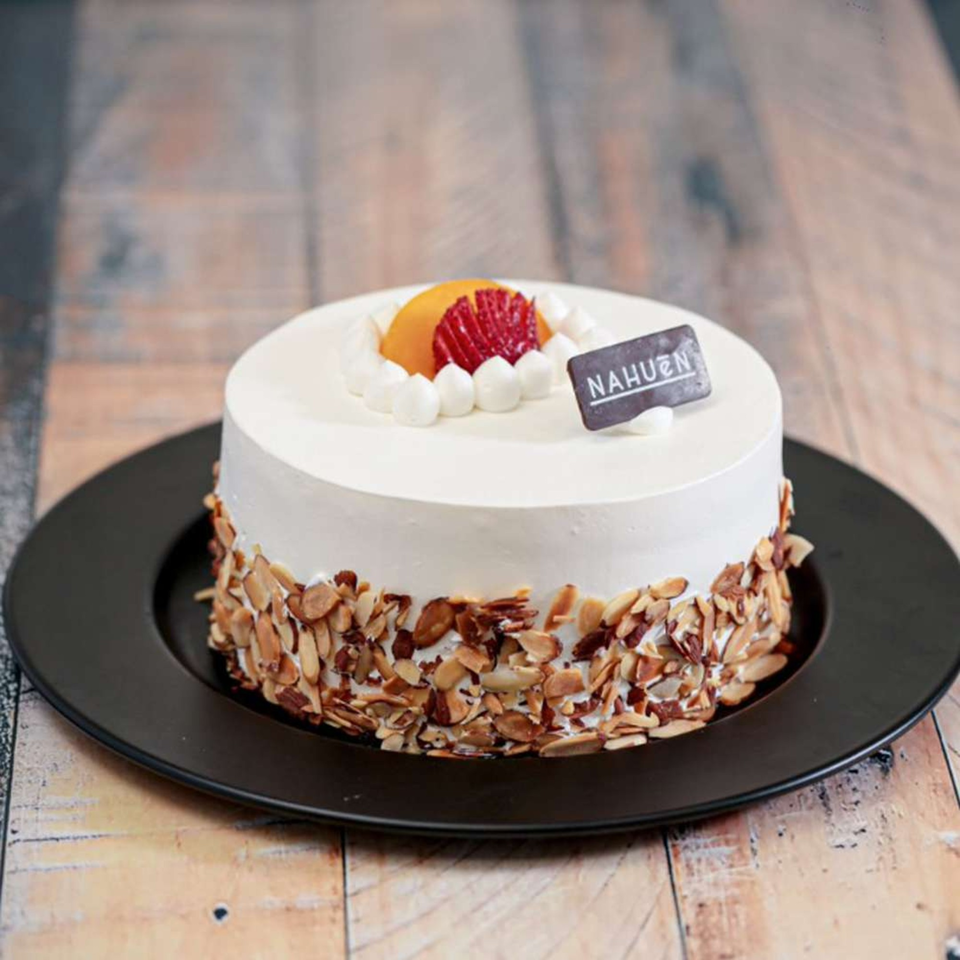 NAHUEN Gourmet Market cake with white frosting, sliced almonds, and fruit on a black plate.